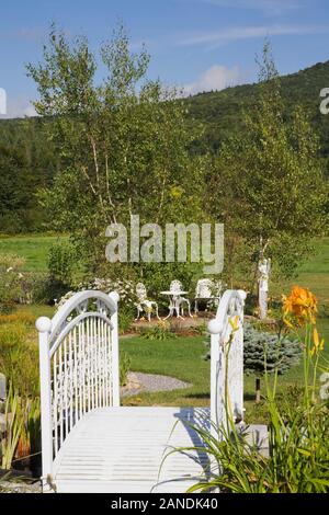 Weiße schmiedeeiserne Steg umgeben von orange Hemerocallis - Daylily Blumen und im Viktorianischen Stil, Tisch, Stühlen im Hinterhof Country Garden. Stockfoto