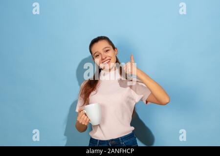 Getränke Kaffee, Tee, zeigt Daumen hoch. Der kaukasischen Teen Girl portrait auf blauem Hintergrund. Schönes Modell in Casual Wear. Konzept der menschlichen Gefühle, Mimik, Vertrieb, Ad. Copyspace. Sieht glücklich aus. Stockfoto