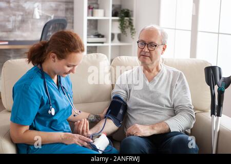 Frau Doktor Lesung Blutdruck von alten Menschen in Pflegeheimen mit digitalen Gerät. Krankenschwester und Patient sitzen auf der Couch. Stockfoto