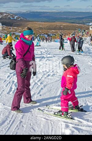 Fröhliche lächelnde Mutter, die kleine Mädchen auf Skiern betrachtet, Cairngorm Mountain Ski Center, Cairngorms National Park, Scottish Highlands, in der Nähe von Aviemore, Großbritannien Stockfoto