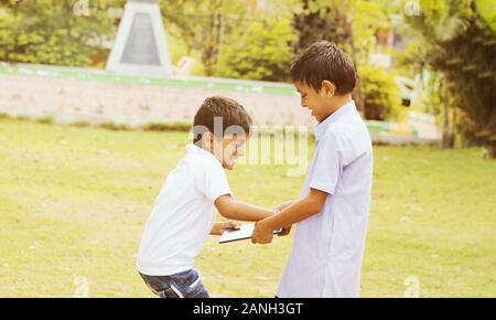 Zwei Kinder kämpfen für Tablet im Park im Freien - Liite Junge versucht, eine Tablette von ihrem Freund oder Bruder zu ergreifen - Begriff der Sucht der Kinder Stockfoto