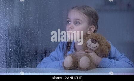 Kleines Mädchen umarmt Teddybären und schauen durch das Fenster im regnerischen Wetter Stockfoto