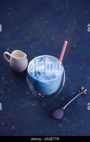 Ein Glas kaltes blau Matcha Tee mit Eis mit Strohhalm, einen Löffel Kaffee Pulver und Milchmann auf einem blauen Tisch. Farbe des Jahres 2020 Classic blau Konzept. Stockfoto
