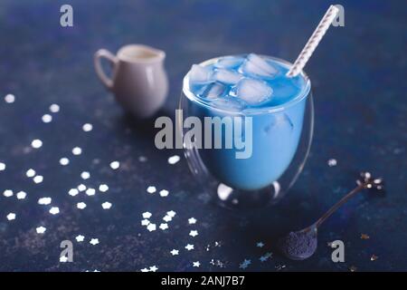 Ein Glas kaltes blau Matcha Tee mit Eis mit Strohhalm, einen Löffel Kaffee Pulver und Milchmann auf einem blauen Tisch. Farbe des Jahres 2020 Classic blau Konzept. Stockfoto