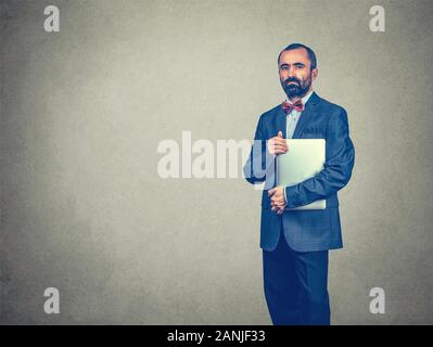 Selbstbewusste, elegante stehend, Bart Mann mit Laptop freuen uns auf Kamera auf grauem Hintergrund. Kurze Haare, kaukasische im dunklen Anzug, Stockfoto
