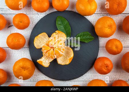 Geöffnet Mandarine mit Blättern auf runde, schwarze Platte mit den ganzen hellen orange Mandarinen auf Risse weiße Holztisch umgeben. Früchte haben natürliche Stockfoto