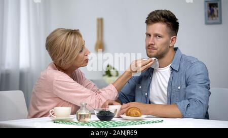 Mutter abwischen gereizte erwachsene Söhne Mund während in Kaffee zu Hause, Pflege Stockfoto
