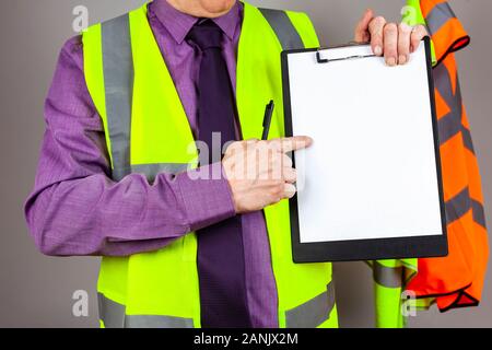 Gesundheit und Sicherheit Ausbilder in einem gelben hohe Sichtbarkeit Weste auf ein leeres weißes Blatt Papier auf einem schwarzen Zwischenablage zeigen Stockfoto