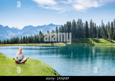 Fröhliche junge Frau sitzt in der Nähe von Lakeside Yoga gegen bewölkter Himmel Stockfoto