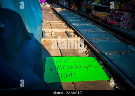 Mae Klong/Thailand-08December2019: Bahnhof Mae Klong mit Gleisen und Zug, die durch fahren, und einem Schild mit der Aufschrift "Zug wird Sie abstürzen" Stockfoto