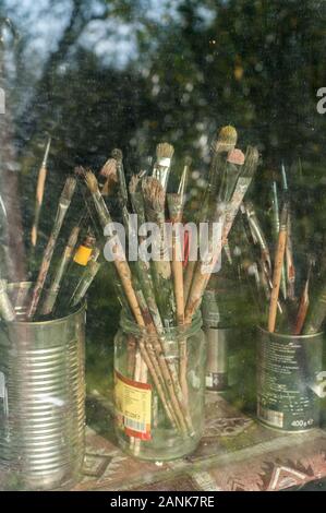 Haufen alter verwendet grunge Pinsel in Dosen und Gläsern hinter Fensterglas in Artist Studio Stockfoto