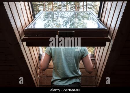 Zurück von einem Mann, der aus einem offenen Fenster auf dem Dach lehnte, im Dachboden, auf dem Glas die Bäume reflektiert werden. Stockfoto