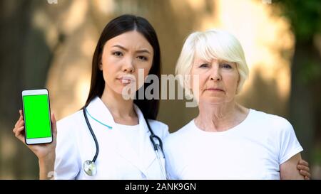 Ernsthafte Krankenschwester mit alten Patienten zeigt Smartphone mit Green Screen an Kamera Stockfoto