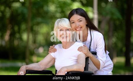 Lächelnd weibliche Krankenschwester umarmen behinderte ältere Frau und Kamera Stockfoto