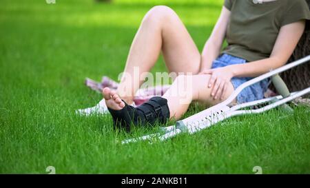 Junge Frau mit den Knöchel verletzt, die Strebe und Krücken sitting park Gras Stockfoto