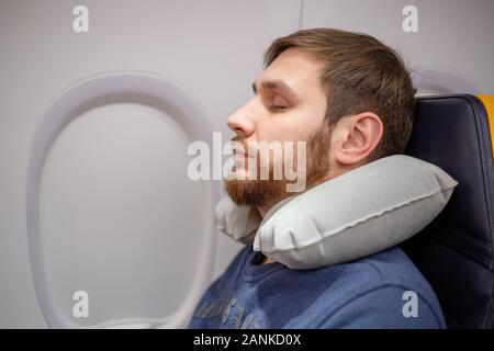 Junger attraktiver europäischer Mann 30 Jahre mit einem Bart schlafen, mit aufblasbarem Nackenkissen im Flugzeug ruhen. Komfort, Stress im Flugzeug Stockfoto