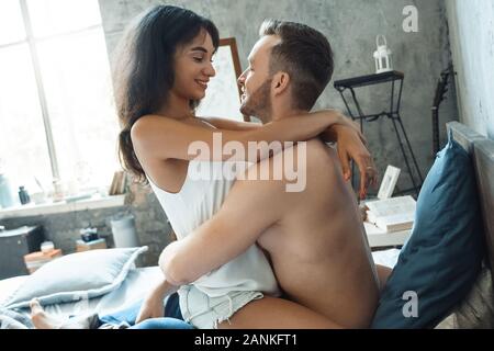 Mixed Race Paar. Junge Frau sitzt auf der Mann auf dem Bett zu Hause sich einander Fröhlich Stockfoto
