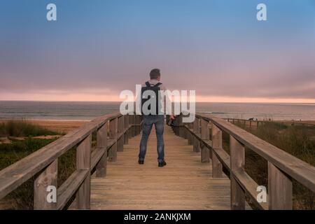 Rückansicht eines Fotografen mit einem Rucksack auf einem Holzsteg mit Blick auf den Ozean. Stockfoto