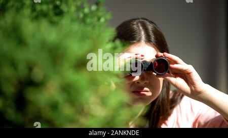 Eifersüchtige Frau mit Fernglas versteckt sich hinter Baum Spionage für Ehemann, Verrat Stockfoto