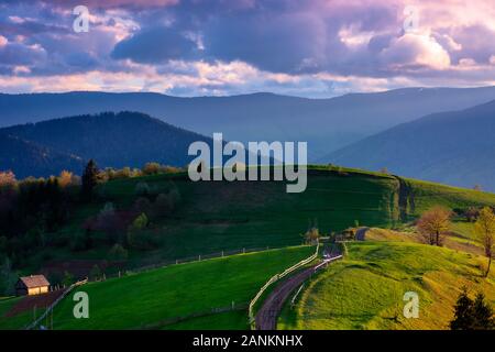 Bergige Landschaft im Abendlicht. hölzernen Zaun entlang dem Weg durch sanfte Hügel, frische grüne Gras. schöne Landschaft im Frühling. Stockfoto