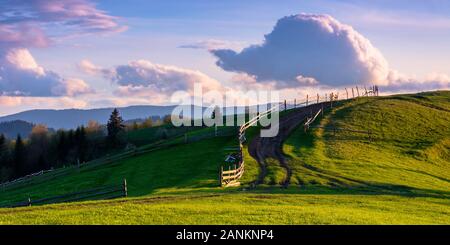 Bergige Landschaft im Abendlicht. hölzernen Zaun entlang dem Weg durch sanfte Hügel, frische grüne Gras. schöne Landschaft im Frühling. Stockfoto