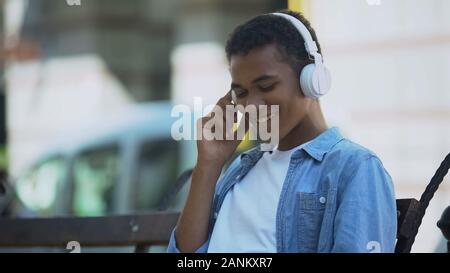Positive junge Mann Hören von Musik über Kopfhörer, sitzt auf der Bank im Freien Stockfoto