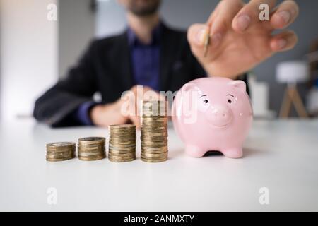 Person's Hand einfügen Münze in das Sparschwein mit zunehmender Münzen Stapel auf dem Schreibtisch Stockfoto