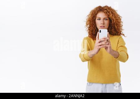 Konzentriert, Ernst - auf der Suche rothaarige curly Frau in gelb Pullover schielen, Blick auf Smartphone Anzeige als Video aufzeichnen ermittelt oder versuchen gute s Stockfoto