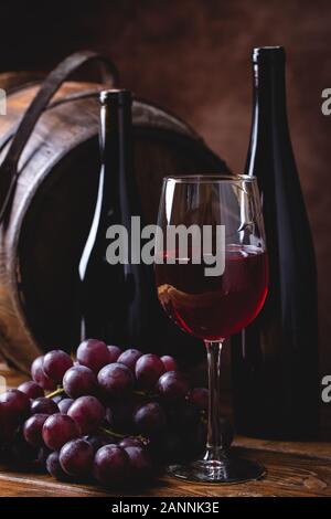 Glas Rotwein und Trauben mit zwei Weinflaschen und Holzfass im Hintergrund Stockfoto