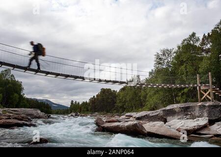 Wanderer mit einem Rucksack, der im Sommer auf der Hängebrücke für Fußgänger den Gebirgsfluss überquert, Norwegen Stockfoto