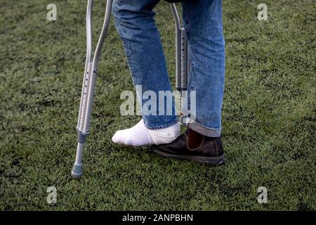 Ein Mann mit einem gebrochenen Bein steht auf Krücken auf dem Rasen eines Fußballfeldes zu einem Stadion Stockfoto