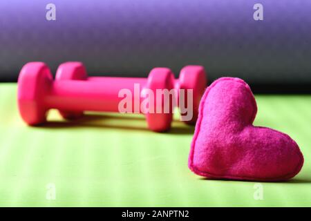 Begeisterung für den Sport und Training. Weibliche Stil und Training Konzept. Hanteln in rosa Farbe neben Soft Toy Herz auf Grün und Lila defokussiertem Hintergrund, aus der Nähe. Herzen vor Barbells auf Yoga Matte Stockfoto