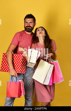 Paar in der Liebe hält Einkaufstaschen auf gelben Hintergrund. Shopping und Ausgaben Konzept. Mann mit Bart hält Red polka gestrichelten Kasten. Kerl mit Bart und Mädchen mit glücklichen Gesichtern einkaufen. Stockfoto