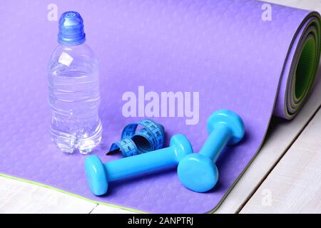Gestaltung und Fitnessgeräte. Training und Sport Konzept. Barbells in der Nähe von Wasser Flasche und cyan Maßband Rollen liegen auf lila Yogamatte. Hanteln aus blauem Kunststoff auf hellen Hintergrund Stockfoto
