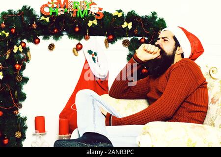 Gut aussehender bärtiger Mann mit langem Bart in Christmas Red Hat und Pullover sitzen und Gähnen auf beige Vintage Sessel um Weihnachten Dekoration, auf weissem Studiohintergrund Stockfoto