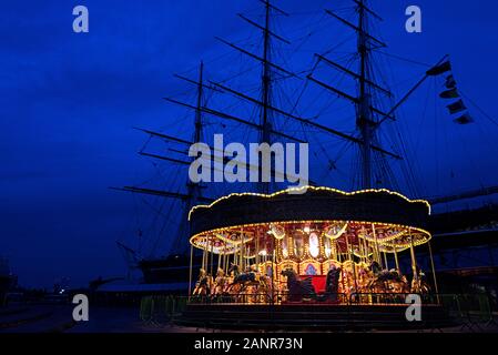 Das Karussell in der Nähe der Cutty Sark leuchtet im Winter Dämmerung, mit der Cutty Sark silhouetted Hinter; Greenwich, London. Stockfoto