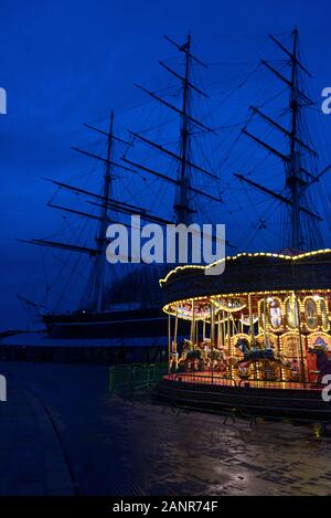 Das Karussell in der Nähe der Cutty Sark leuchtet im Winter Dämmerung, mit der Cutty Sark silhouetted Hinter; Greenwich, London. Stockfoto