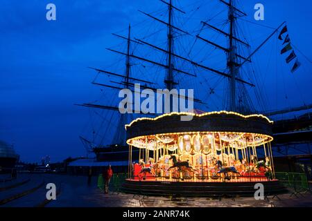 Das Karussell in der Nähe der Cutty Sark leuchtet im Winter Dämmerung, mit der Cutty Sark silhouetted Hinter; Greenwich, London. Stockfoto