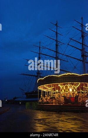 Das Karussell in der Nähe der Cutty Sark leuchtet im Winter Dämmerung, mit der Cutty Sark silhouetted Hinter; Greenwich, London. Stockfoto