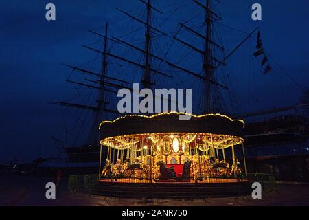 Das Karussell in der Nähe der Cutty Sark leuchtet im Winter Dämmerung, mit der Cutty Sark silhouetted Hinter; Greenwich, London. Stockfoto