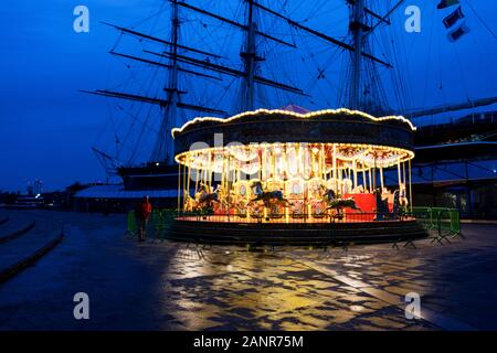 Das Karussell in der Nähe der Cutty Sark leuchtet im Winter Dämmerung, mit der Cutty Sark silhouetted Hinter; Greenwich, London. Stockfoto