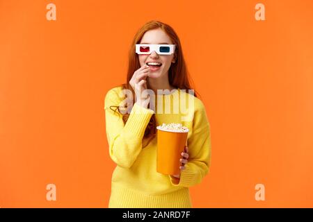 Interessiert und amüsiert lächelnd geeky rothaarige Frau in gelb Pullover, Film in Kino mit 3D-Brille, essen Popcorn und starren neugierig Stockfoto