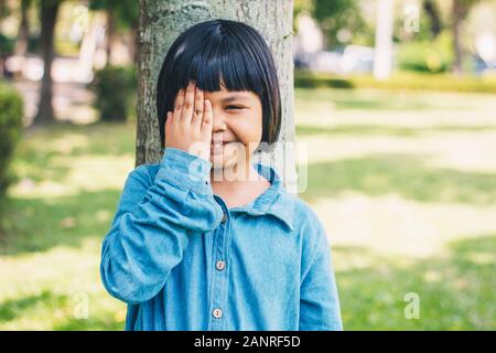 Das Bild von einem kleinen Mädchen mit ihren Händen, ihrem rechten Auge gegen einen Baum im Park gelehnt. Stockfoto