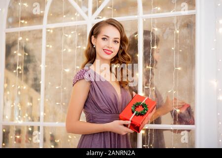 Schöne Mädchen mit roten Lippen und Make-up hält Geschenk auf dem Hintergrund von Bokeh und Weihnachtsbaum Stockfoto