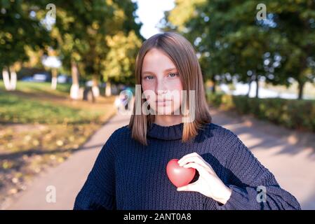 Ein jugendlicher Mädchen zwischen 12 und 15 Jahren, die im Sommer in der Stadt, hält ein Spielzeug, das Herz in der Hand, Konzept Geschenk Liebe parenting Hilfe und Support. Aus Gewirken Stockfoto