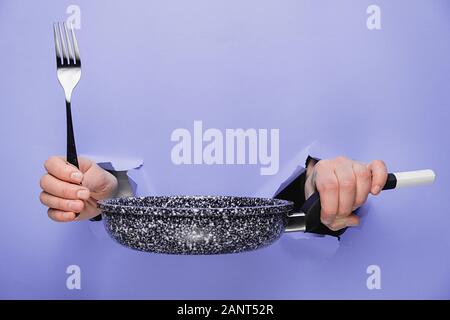 In der Nähe der weiblichen Händen hält Pfanne und durch Papierreste lila Hintergrund Gabel. Küchengeräte Konzept. Kopieren Sie Platz für Werbung Stockfoto