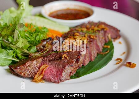 Schneiden Sie mittelgroßes, rares gegrilltes Rindersteak mit gebratenem Knoblauch mit thailändischer, würziger Soße und Gemüse auf der Seite. Stockfoto