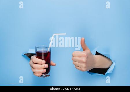 Weibliche hand mit einem Glas frischen roten Saft mit einem Strohhalm durch zerrissene blaues Papier an der Wand. Fruchtsäfte und gesunde Ernährung. Handzeichen. Loch in der Wand. Stockfoto