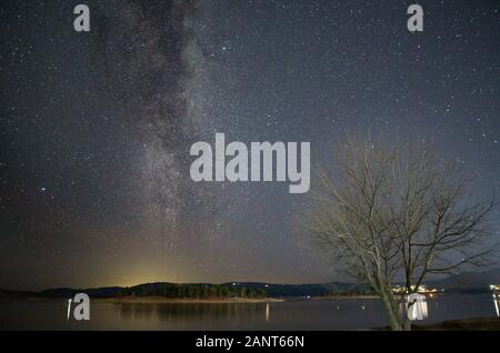 Milchstraße über einen blattlosen Baum am Ufer des Lake Jindabyne Jindabyne mit der Stadt im Hintergrund Stockfoto