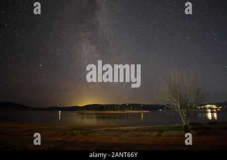 Milchstraße über einen blattlosen Baum am Ufer des Lake Jindabyne Jindabyne mit der Stadt im Hintergrund Stockfoto